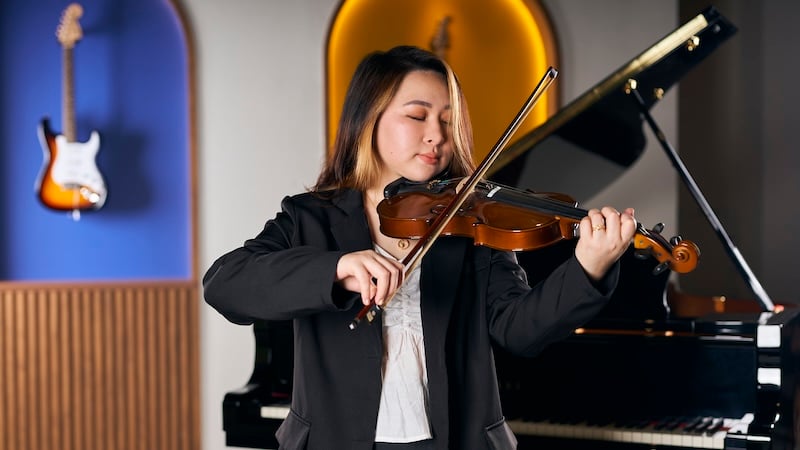A violinist passionately playing the violin with her eyes closed. She is in a music studio with a grand piano in the background.
