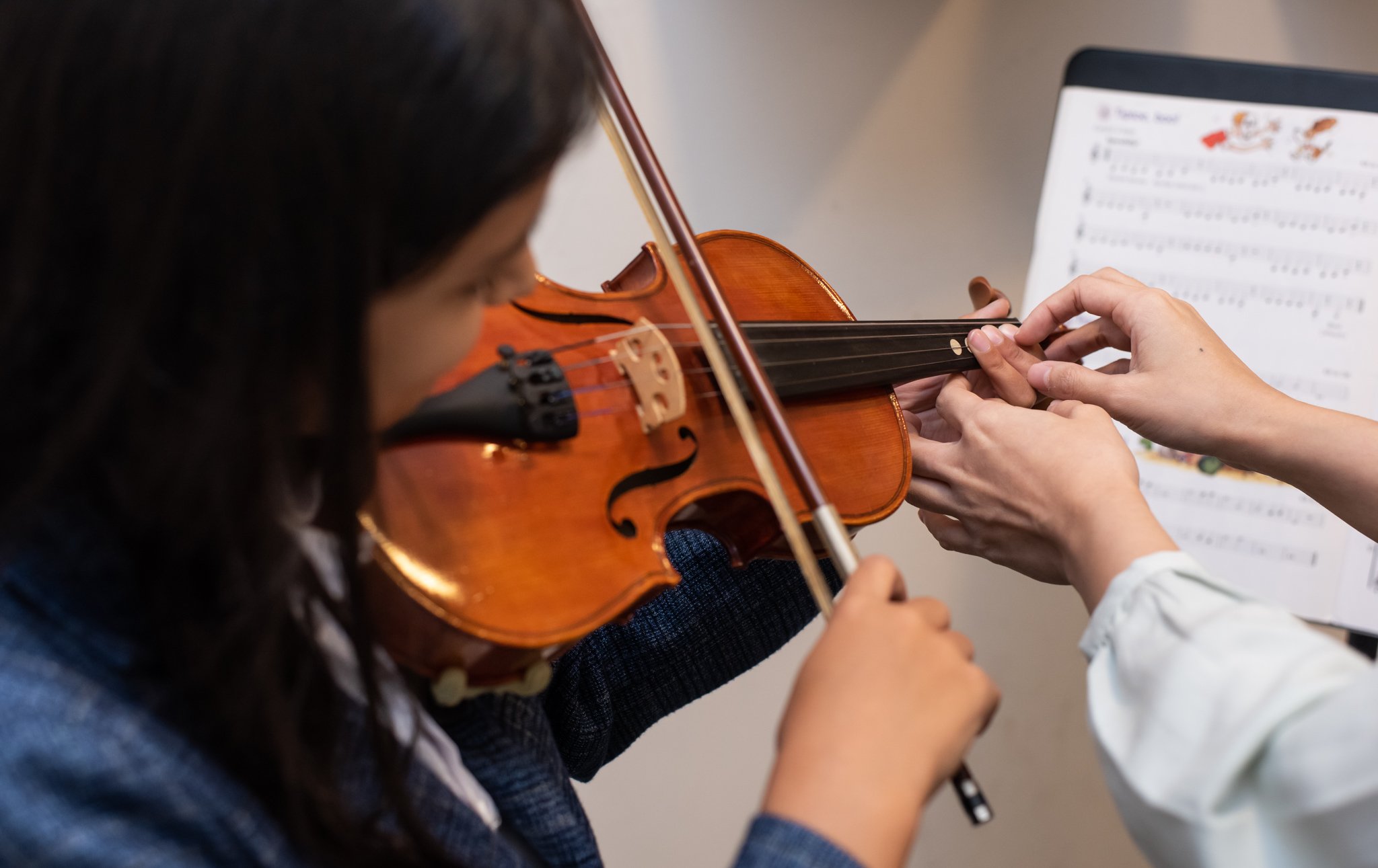A student is holding a violin and an off-screen teacher is correcting her finger placement on the strings.