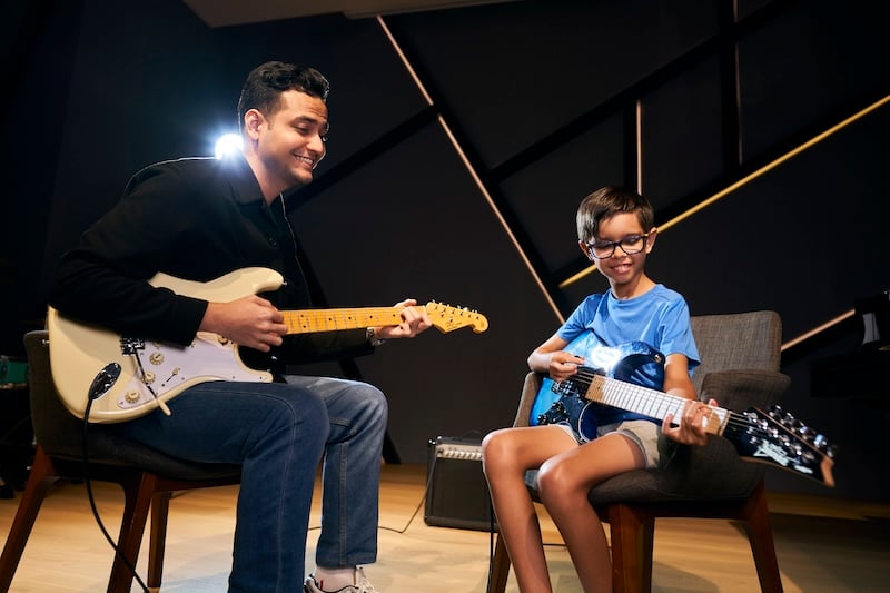 A music teacher holding an electric guitar is seated across from his student who is also holding an electric guitar. They are both smiling. 